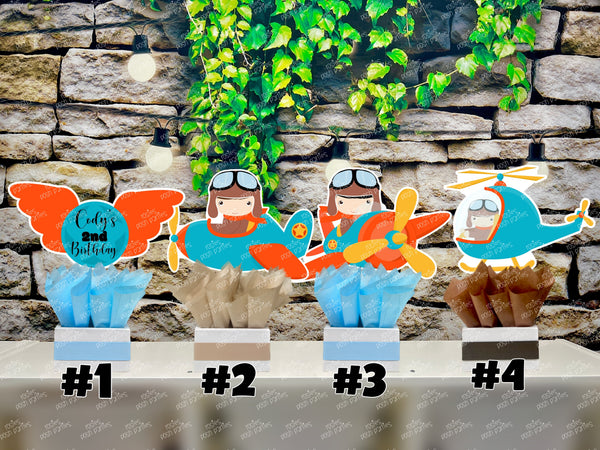 Set of boy airplane pilot-themed birthday party table centerpieces from Rosie's Posh Parties. Features a personalized sign with orange wings, two boy pilots flying blue and orange airplanes, and a pilot flying a blue helicopter. The cutouts are nestled in square bases decorated with blue, tan, and brown tissue paper.