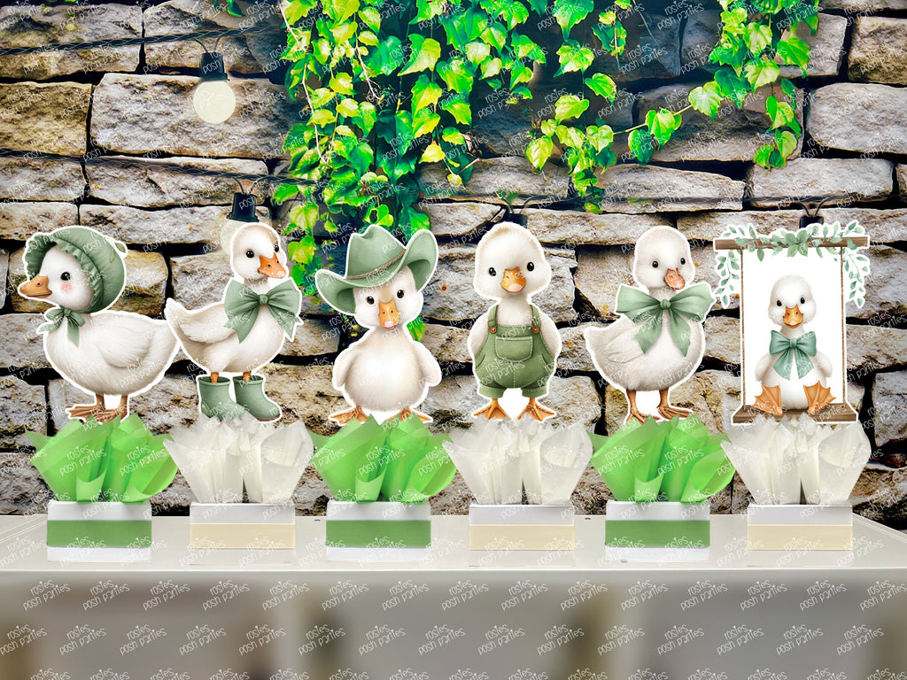 fully assembled party centerpieces featuring adorable watercolor-style white baby geese or ducklings dressed in pink outfits. The designs include Goose w/ Bonnet, Goose w/ Boots, Goose w/Cowboy Hat, Goose w/ Overalls, Goose Standing, Goose on Swing. Each cutout stands on a white base wrapped in green ribbon with fluffy green tissue paper