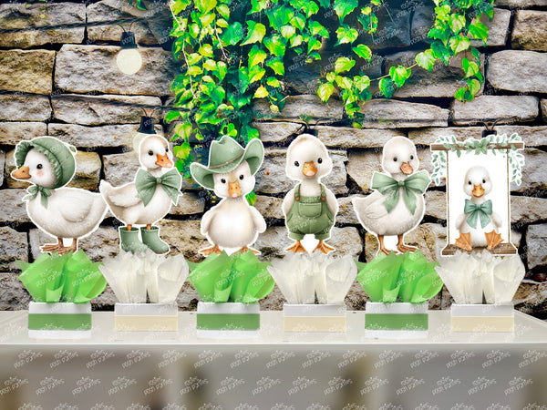 fully assembled party centerpieces featuring adorable watercolor-style white baby geese or ducklings dressed in pink outfits. The designs include Goose w/ Bonnet, Goose w/ Boots, Goose w/Cowboy Hat, Goose w/ Overalls, Goose Standing, Goose on Swing. Each cutout stands on a white base wrapped in green ribbon with fluffy green tissue paper