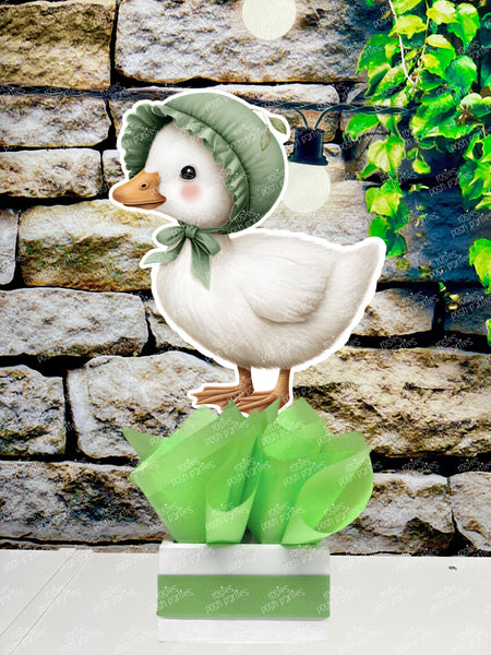fully assembled party centerpieces featuring adorable watercolor-style white baby geese or ducklings dressed in pink outfits. The designs include Goose w/ Bonnet, Goose w/ Boots, Goose w/Cowboy Hat, Goose w/ Overalls, Goose Standing, Goose on Swing. Each cutout stands on a white base wrapped in green ribbon with fluffy green tissue paper