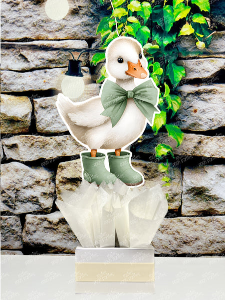 fully assembled party centerpieces featuring adorable watercolor-style white baby geese or ducklings dressed in pink outfits. The designs include Goose w/ Bonnet, Goose w/ Boots, Goose w/Cowboy Hat, Goose w/ Overalls, Goose Standing, Goose on Swing. Each cutout stands on a white base wrapped in green ribbon with fluffy green tissue paper