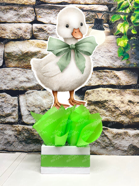 fully assembled party centerpieces featuring adorable watercolor-style white baby geese or ducklings dressed in pink outfits. The designs include Goose w/ Bonnet, Goose w/ Boots, Goose w/Cowboy Hat, Goose w/ Overalls, Goose Standing, Goose on Swing. Each cutout stands on a white base wrapped in green ribbon with fluffy green tissue paper