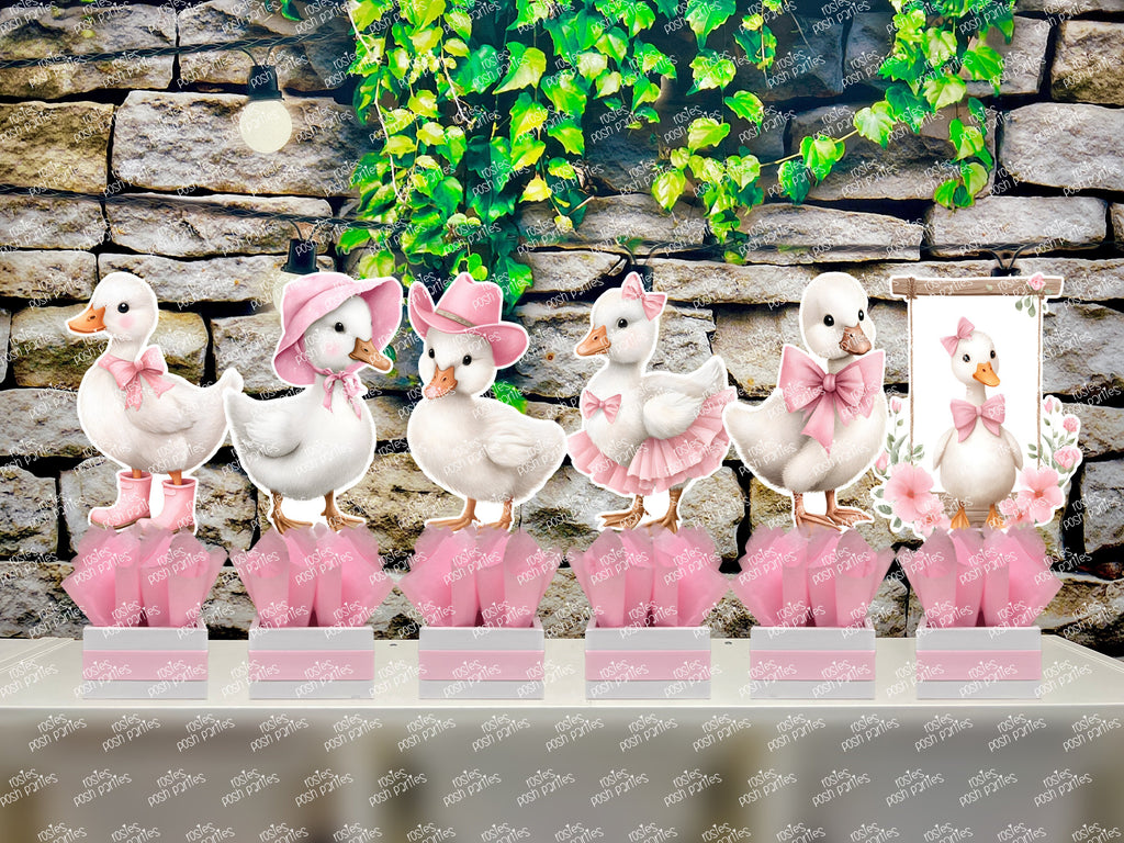Set of 6 fully assembled party centerpieces featuring adorable watercolor-style white baby geese or ducklings dressed in pink outfits. The designs include a goose in pink boots, a pink sunhat, a pink cowboy hat, a pink tutu, a large pink bow, and one inside a pink floral frame. Each cutout stands on a white base wrapped in pink ribbon with fluffy pink tissue paper.