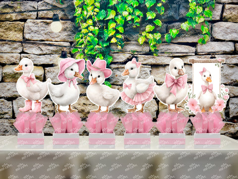 Set of 6 fully assembled party centerpieces featuring adorable watercolor-style white baby geese or ducklings dressed in pink outfits. The designs include a goose in pink boots, a pink sunhat, a pink cowboy hat, a pink tutu, a large pink bow, and one inside a pink floral frame. Each cutout stands on a white base wrapped in pink ribbon with fluffy pink tissue paper.