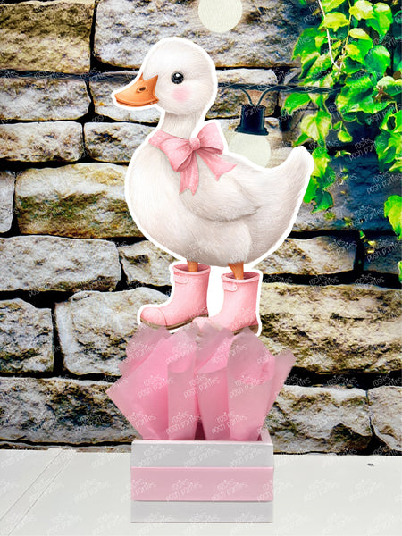 fully assembled party centerpieces featuring adorable watercolor-style white baby geese or ducklings dressed in pink outfits. The designs include a goose in pink boots, a pink sunhat, a pink cowboy hat, a pink tutu, a large pink bow, and one inside a pink floral frame. Each cutout stands on a white base wrapped in pink ribbon with fluffy pink tissue paper.