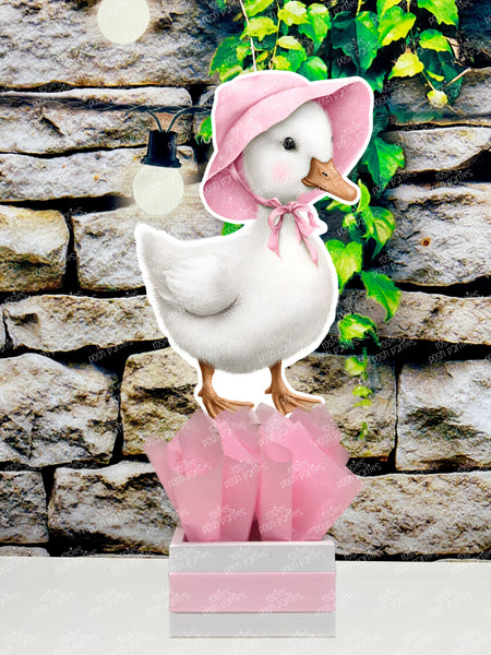 fully assembled party centerpieces featuring adorable watercolor-style white baby geese or ducklings dressed in pink outfits. The designs include a goose in pink boots, a pink sunhat, a pink cowboy hat, a pink tutu, a large pink bow, and one inside a pink floral frame. Each cutout stands on a white base wrapped in pink ribbon with fluffy pink tissue paper.