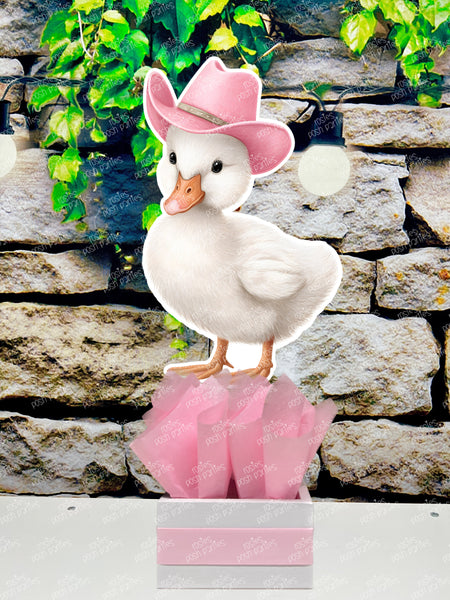 fully assembled party centerpieces featuring adorable watercolor-style white baby geese or ducklings dressed in pink outfits. The designs include a goose in pink boots, a pink sunhat, a pink cowboy hat, a pink tutu, a large pink bow, and one inside a pink floral frame. Each cutout stands on a white base wrapped in pink ribbon with fluffy pink tissue paper.
