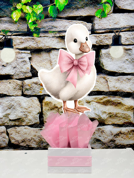 fully assembled party centerpieces featuring adorable watercolor-style white baby geese or ducklings dressed in pink outfits. The designs include a goose in pink boots, a pink sunhat, a pink cowboy hat, a pink tutu, a large pink bow, and one inside a pink floral frame. Each cutout stands on a white base wrapped in pink ribbon with fluffy pink tissue paper.