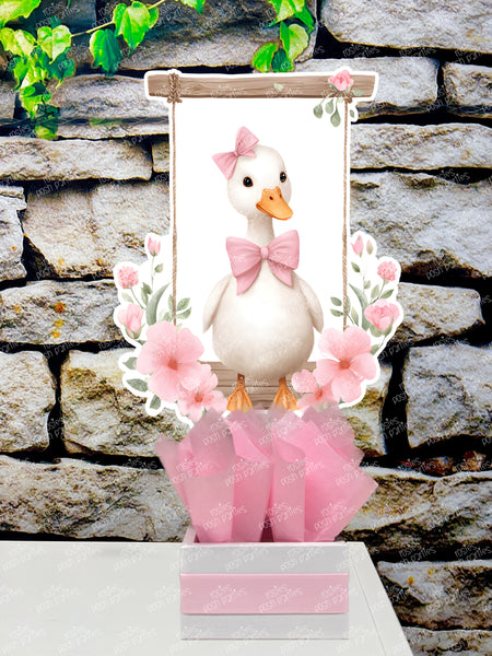 fully assembled party centerpieces featuring adorable watercolor-style white baby geese or ducklings dressed in pink outfits. The designs include a goose in pink boots, a pink sunhat, a pink cowboy hat, a pink tutu, a large pink bow, and one inside a pink floral frame. Each cutout stands on a white base wrapped in pink ribbon with fluffy pink tissue paper.